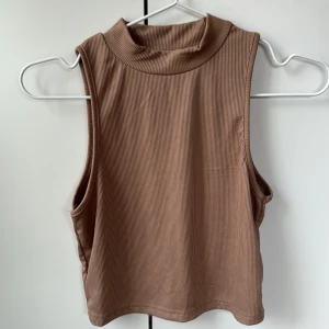 Beige ribbad ärmlös polotopp - Säljer en stilren beige ribbad polotopp utan ärmar. Perfekt för lager-på-lager eller som den är. Den har en hög krage som ger en elegant touch. Passar både till vardags och mer uppklädda tillfällen.