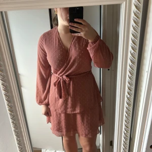 Rosa omlottklänning - Säljer en söt rosa omlottklänning med v-ringning och knytband i midjan. Klänningen har långa ärmar och ett diskret prickigt mönster. Perfekt för en somrig look!