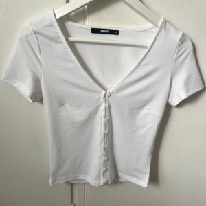 Vit kortärmad topp från Bik Bok❤️ - Säljer en stilren vit topp från Bik Bok med korta ärmar och en v-ringad design. Toppen har knappar framtill och är perfekt för en enkel och fräsch look. Passar bra till både jeans och kjol!