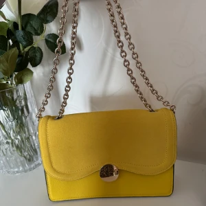 Gul axelväska med kedja - Axelväska med en elegant kedjerem och rund metallknapp. Väskan har en stilren design med en ljus insida och praktiskt fack. Perfekt för att liva upp din outfit! 💛