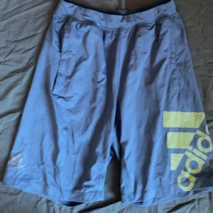 Blå shorts från Adidas - Säljer ett par blå shorts från Adidas med en stor logga i ljusgrönt på benet. Shortsen har en normal passform och är perfekta för träning. De har praktiska fickor på sidorna.