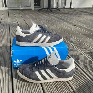 Adidas Gazelle i mörkblå mocka - Snygga Adidas Gazelle sneakers i mörkblå mocka med vita detaljer och klassiska tre ränder på sidan. Skorna har snörning och en bekväm passform, perfekt för en stilren look.