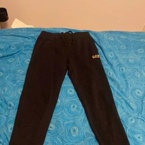 Svarta mjukisbyxor från Emporio Armani - Snygga svarta mjukisbyxor från Emporio Armani med EA7-logga i guld. Byxorna har en bekväm passform med snörning i midjan och är perfekta för en avslappnad stil. Säljer pga för små sparsamt använda.