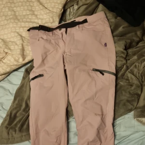 Rosa "raggarbyxor" från Trekmates - Säljer ett par rosa "raggar" byxor från Trekmates i storlek XS. Byxorna har praktiska dragkedjor på benen och en justerbar midja med svarta detaljer. Perfekta för utomhusaktiviteter och äventyr.