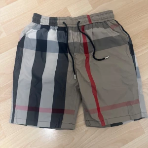 Badshorts - Snygga badshorts med ett klassiskt rutigt mönster i beige, svart och rött. De har en elastisk midja med snörning för justerbar passform. Perfekta för sommarens stranddagar!