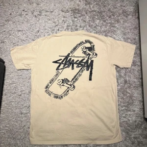 Beige t-shirt från Stussy - Säljer en beige t-shirt från Stussy med ett stort tryck på baksidan och en mindre logga på framsidan. T-shirten har korta ärmar och är perfekt för en avslappnad stil. Passar bra till både jeans och shorts.