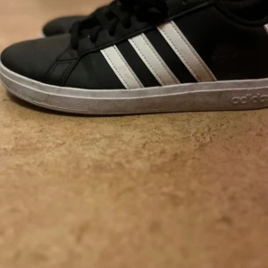 Svarta Adidas sneakers med vita ränder - Säljer ett par klassiska svarta Adidas sneakers med de ikoniska tre vita ränderna på sidan. Skorna har en rund tå och snörning framtill. Perfekta för en avslappnad stil.