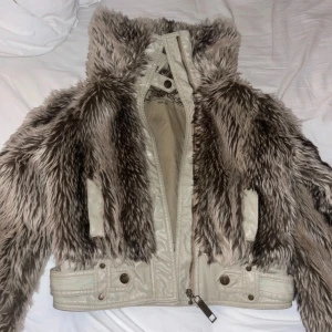 Beige jacka med fuskpäls - Säljer en unik beige jacka från Miss Sixty med fuskpälsdetaljer. Jackan har en dragkedja framtill och knappar vid kragen. Den är fodrad och har en cool, edgy look. Perfekt för att sticka ut i mängden!