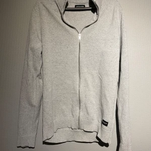 Grå kofta från Jack & Jones - Säljer en stilren grå kofta från Jack & Jones med dragkedja framtill. Koftan har långa ärmar och en ribbad kant vid ärmslut och nederkant. Perfekt för en avslappnad look.