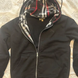 Svart hoodie från Burberry med dragkedja - Svart hoodie från Burberry med klassiskt rutigt foder i huvan och dragkedja framtill. Tröjan har två stora fickor och diskret broderad logga på bröstet. Perfekt för dig som gillar stilrena och exklusiva plagg. Helt ny