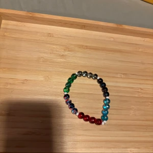 Färgglatt pärlarmband i glas - Ett armband med glaspärlor i olika färger som rött, grönt, blått och silver. Pärlorna är runda och sitter på en elastisk tråd, vilket gör det enkelt att ta på och av. Perfekt accessoar för att addera lite färg till din stil.