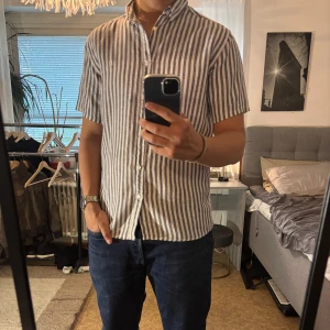 Randig kortärmad skjorta Les Deux - Snygg kortärmad skjorta från Les Deux i vitt och mörkblått randigt mönster. Skjortan har klassisk krage, knäppning framtill och är tillverkad i luftig linneblandning – perfekt för varma dagar. Stilren och enkel att matcha med jeans eller shorts.