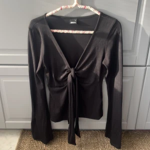 Svart omlottopp med knyt från Gina Tricot - Svart långärmad omlottopp från Gina Tricot i mjuk bomullsmix. Toppen har djup v-ringning och snygg knytning framtill som ger en trendig look. Perfekt att styla med jeans eller kjol för en cool vibe.