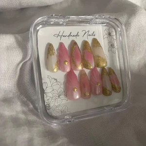 Handgjorda rosa och guldiga lösnaglar - Säljer ett set med handgjorda lösnaglar i mandelform. Naglarna har en mix av rosa och nude bas med marmorerade detaljer i guld och små guldfärgade dekorationer. Kommer i en genomskinlig ask och inkluderar lim, nagelfil och träpinne för applicering.