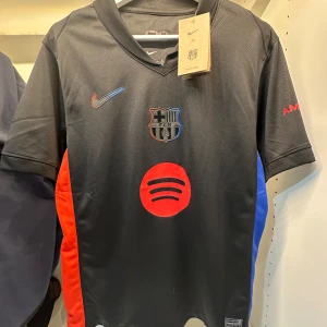 Svart FC Barcelona Nike fotbollströja - Snygg svart FC Barcelona matchtröja från Nike med klubbmärke på bröstet, röd och blå Nike-logga och röd Spotify-logga på magen. Tröjan har korta ärmar, V-ringning och paneler i rött och blått på sidorna. Tillverkad i lätt och ventilerande Dri-FIT-material.