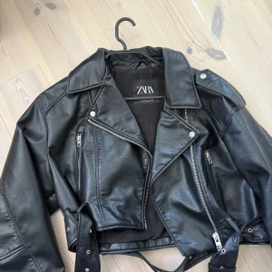 Svart skinnjacka från Zara XL - Snygg svart skinnjacka från Zara i bikerstil med coola silverfärgade dragkedjor, bred krage och bälte i midjan. Jackan har långa ärmar och detaljer som axelklaffar och flera fickor med dragkedja. Perfekt för dig som vill ha en edgy look.