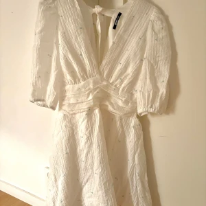 Vit omlottklänning med knyt i ryggen - Superfin vit kortklänning  med omlottliv, knyt i ryggen och diskreta små blå broderade detaljer. Klänningen har puffiga trekvartsärmar och är gjord i ett luftigt bomullstyg, perfekt för varma dagar.