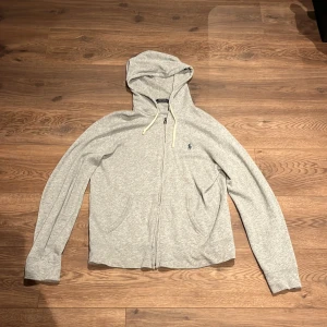 Grå hoodie från Polo Ralph Lauren - Snygg gråmelerad hoodie från Polo Ralph Lauren med dragkedja och vit snörning i huvan. Klassisk blå logga broderad på bröstet och två fickor framtill. Mjuk bomullskänsla och långärmad modell, perfekt för chill dagar.