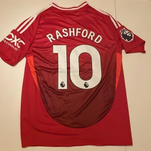 Manchester United Rashford 10 tröja - Snygg Manchester United matchtröja replika med Rashford och nummer 10 på ryggen. Röd med vita detaljer, Premier League-logga på ärmen och sponsortryck på bröstet. Tillverkad i lätt och ventilerande polyester, klassisk Adidas-design med tre vita ränder på axlarna. Handtvätt endast!