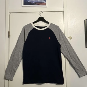 Blå långärmad tröja Polo Ralph Lauren - Snygg långärmad tröja från Polo Ralph Lauren i marinblått med grå ärmar och vit krage. Framsidan har en liten röd logga och baksidan har en stor vit 67-tryck. Tillverkad i 100% bomull, perfekt för en sportig och avslappnad look.
