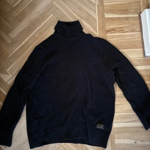 Svart ribbad polotröja - Svart polotröja med ribbad struktur och hög krage. Tröjan har lång ärm och en liten diskret etikett nertill. Perfekt för lager-på-lager och passar till både jeans och kjol.