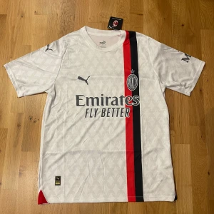 AC Milan vit bortatröja 23/24 Puma L - AC Milan bortatröja från Puma i vitt med subtilt mönster, röd och svart rand längs framsidan. Klassisk rund hals, korta ärmar och tryckta loggor från Emirates och Wefox. Materialet är lätt och andas, perfekt för match eller träning.