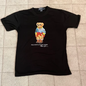 Svart Polo Bear t-shirt från Ralph Lauren - Svart t-shirt från Polo Ralph Lauren med ikoniskt Polo Bear-tryck på bröstet. Klassisk rund hals och korta ärmar. Tillverkad i 100% bomull för en skön och avslappnad känsla. Perfekt för dig som gillar streetstyle och vill sticka ut med en cool print. Passar även S