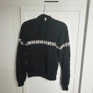 Svart hoodie med vit text från NA-KD - Cool svart hoodie från NA-KD med vit grafisk text runt hela bröstet och armarna. Tröjan har huva med snörning och en stor magficka framtill. Perfekt för en avslappnad och trendig look.