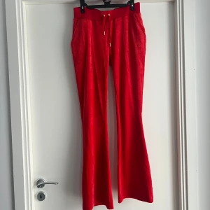 Röda velourbyxor Juicy Couture S - Röda mjukisbyxor från Juicy Couture i velour med bootcut-ben och snörning i midjan. Byxorna har broderad JC-logga framtill och elastisk midja för skön passform. Perfekta för en chill och trendig look.