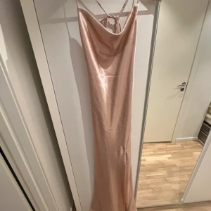 Champagnefärgad långklänning från True Decadence - Snygg långklänning i skimrande champagnefärg från True Decadence. Klänningen har tunna axelband, öppen rygg med snörning och ett elegant fall. Materialet är glansigt och känns lyxigt, perfekt för speciella tillfällen. Aldrig använd