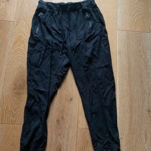 Svarta mjukisbyxor från Happy Holly - Svarta mjukisbyxor från Happy Holly med dragsko i midjan och dragkedjor på fickorna. Byxorna har en relaxed fit och är gjorda i mjukt material, perfekta för chill dagar. Elastisk midja för extra komfort.