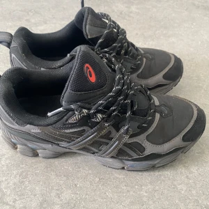 Svarta Asics GEL sneakers - Svarta Asics GEL sneakers med grå detaljer och reflekterande partier. Skorna har snörning med svarta och vita skosnören, samt röd Asics-logga på plösen och GEL-märkning bak. Ovandelen är i syntet och mesh för bra andningsförmåga och komfort.