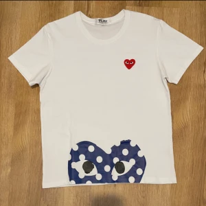 Vit Comme des Garçons Play t-shirt - Vit t-shirt från Comme des Garçons Play med klassiskt rött hjärta på bröstet och stor blå prickig hjärtdetalj med ögon längst ner. T-shirten är i bomull och har korta ärmar och rund hals. Perfekt statement-plagg för dig som gillar coola prints. Storleken i tröjan är L men sitter som eu s
