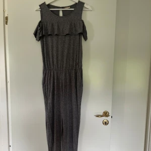Grå jumpsuit med volangdetalj - Snygg grå jumpsuit med volang över axlarna och öppning i ryggen. Mjuk och stretchig känsla, resår i midjan för skön passform. Perfekt för dig som gillar en avslappnad men ändå trendig look.