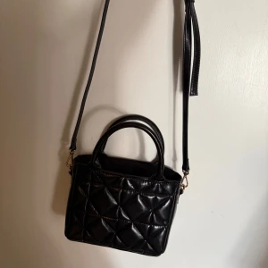Svart quiltad axelväska med handtag - Snygg svart axelväska med quiltad design och två rejäla handtag. Väskan har justerbar axelrem och är tillverkad i glansigt skinnimitation. Perfekt storlek för det viktigaste och ger en trendig vibe till din outfit.