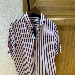 Randig kortärmad skjorta  - Snygg kortärmad skjorta  med vertikala ränder i rött, blått och vitt. Klassisk krage med knappar, bröstficka och relaxed fit. Perfekt för en fräsch och stilren look under varmare dagar.