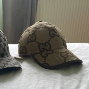 Gucci keps - Snygg beige keps från Gucci med det klassiska GG-monogrammet i brunt över hela kepsen. Justerbar rem baktill i brunt läder och böjd skärm. Tillverkad i slitstarkt canvasmaterial som ger en exklusiv känsla och streetstyle-vibe.