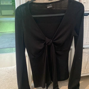 Svart långärmad knytblus Gina Tricot - Svart långärmad topp från Gina Tricot med djup v-ringning och snygg knytning framtill. Tunn och stretchig modell som sitter figurnära och har en stilren look. Perfekt att styla med jeans eller kjol för en trendig outfit.