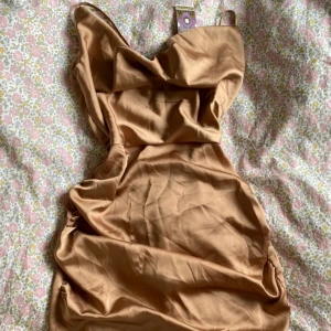 Beige satin minidress med drapering - Superfin beige minidress i satin med draperad urringning och smala axelband. Klänningen har en figurnära passform och en lätt glansig yta som ger en lyxig känsla. Perfekt för dig som vill sticka ut med en elegant och trendig look.