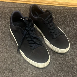 Marinblå sneakers från Selected Homme - Stilrena svarta sneakers från Selected Homme med vit platt sula och klassisk snörning. Ovandelen är i mocka och insidan är vadderad för extra komfort. Perfekta för dig som gillar en clean och enkel look. Använd ett fåtal gånger, skick 9/10. Priset kan diskuteras.