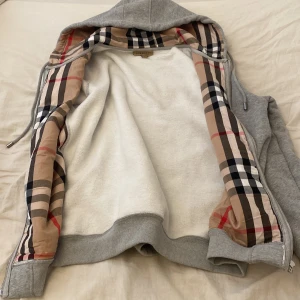 Grå hoodiejacka från Burberry, L - Snygg grå hoodiejacka från Burberry med klassiskt rutigt foder i beige, svart, vitt och rött. Jackan har dragkedja, huva med snörning och broderad Burberry-logga på bröstet. Perfekt för dig som vill ha en stilren och lyxig streetwear-look.