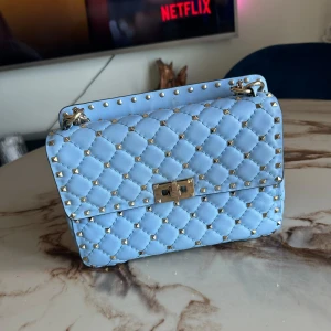 Valentino Rockstud Spike Bag - Säljer en ljusblå Rockstar Spike Bag handväska från Valentino med silverfärgade nitar och kedjedetaljer. Väskan har ett elegant rutmönster, vridlås i metall och är tillverkad i skinn. Perfekt för dig som vill sticka ut med en lyxig accessoar.