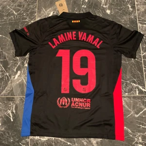 FC Barcelona Lamine Yamal Nike tröja - Snygg svart FC Barcelona fotbollströja från Nike med Lamine Yamal och nummer 19 på ryggen. Tröjan har röda och blå sidopaneler, klubbmärke på bröstet och tryck från Cactus Jack samt UNHCR-logga. Tillverkad i lätt och ventilerande material, perfekt för match eller träning.