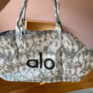 Gråmönstrad shoppingväska från Alo - Stor och rymlig shoppingväska från Alo i grått och vitt mönster. Väskan är tillverkad i 100% bomull och har två rejäla handtag. Snygg logga framtill och coolt abstrakt mönster som sticker ut. Perfekt för att bära med sig allt man behöver till skolan, gymmet eller stan.