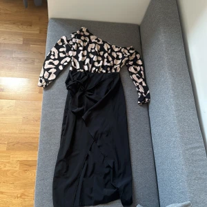 Svart långklänning med leopardmönster - Snygg långklänning med svart kjol och omlott-detalj samt knyt i midjan. Överdelen har beige och svart leopardmönster och långa ärmar med resår vid handlederna. 