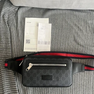 Svart Gucci axelväska med GG-mönster - Säljer en svart axelväska från Gucci med klassiskt GG-mönster och silverfärgad dragkedja. Väskan har en röd och marinblå justerbar rem och detaljer i skinn. Perfekt för att bära det viktigaste med stil.