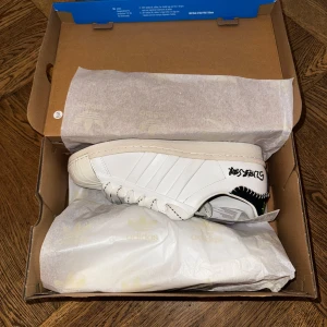 Adidas Superstar x Jonah Hill – strl 41 1/3 - Limitied Edition från Jonah Hills samarbete med Adidas. Ett par stilrena Adidas Superstars med broderade detaljer och tjockare snören i retro-look. Köpta från SNS drop 2020. Kvitto och orderbekräftelse finns.  👟 Skick: Helt oanvända 📏 Storlek: 41 1/3 📦 Levereras i originalkartong. Priset är förhandlingsbart.  