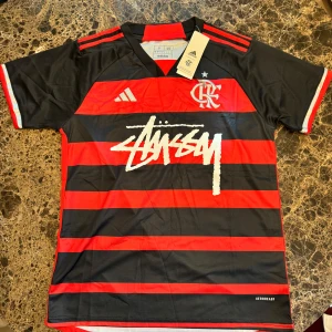 Adidas x Stüssy Flamengo fotbollströja - Snygg Flamengo fotbollströja från Adidas i samarbete med Stüssy. Tröjan är svart och röd med horisontella ränder, korta ärmar och vit Stüssy-logga på bröstet. Klassisk klubbemblem och Adidas-logga framtill. Tillverkad i lätt och ventilerande material.
