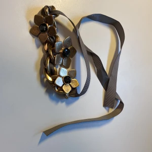 Blommigt guldigt halsband med band *Endast 1 Kvar* - Unikt halsband med stora blommor i guldtonad metall och svarta stenar i mitten. Kedjan är grov och flätad med ett grått band som knyts i nacken. Perfekt statement piece för dig som vill sticka ut med coola detaljer och blommig design.
