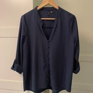 Mörkblå skjorta från Vero Moda, M - Snygg mörkblå skjorta från Vero Moda i storlek M. Skjortan har diskreta ränder, v-ringning och klädda knappar framtill. Långärmad modell med möjlighet att vika upp ärmarna och fästa med knapp. Perfekt för en stilren look.